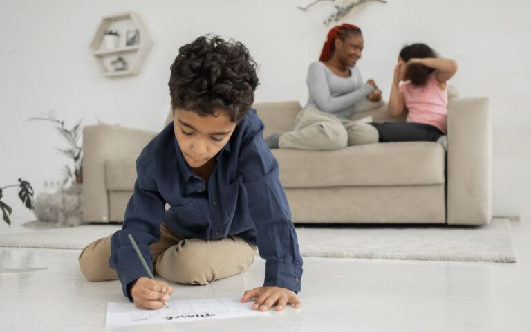 Smart little ethnic boy in casual clothes sitting on floor and solving exercise near young happy mother and sister playi