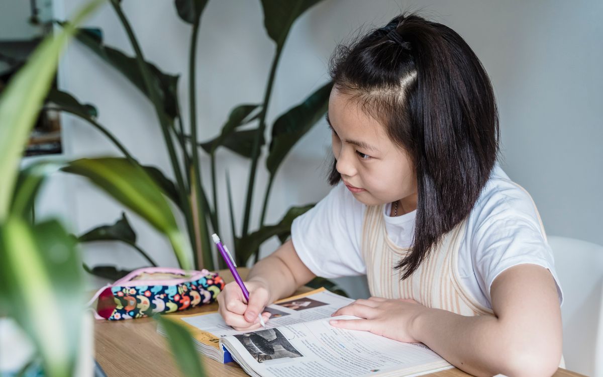 A focused young girl writing in a notebook at a wooden table with indoor plants.