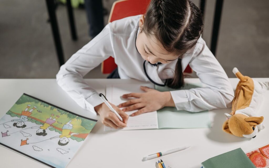 A young girl focused on homework, writing in a notebook in a classroom setting.