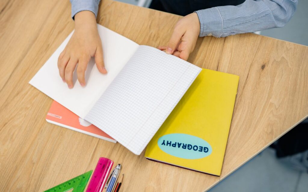 Hands flipping through a geography notebook on a wooden desk with pens and rulers.