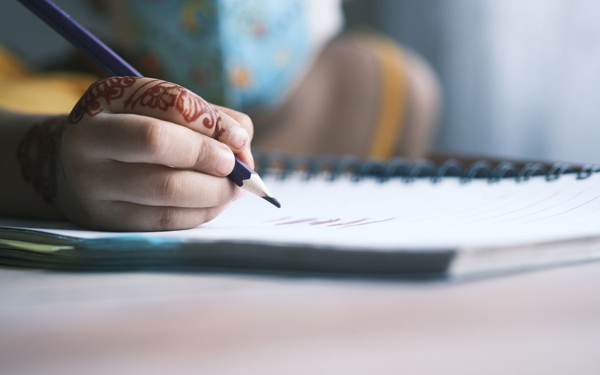 Close-up of a child's hand writing in a notebook with colorful pencils and henna design.