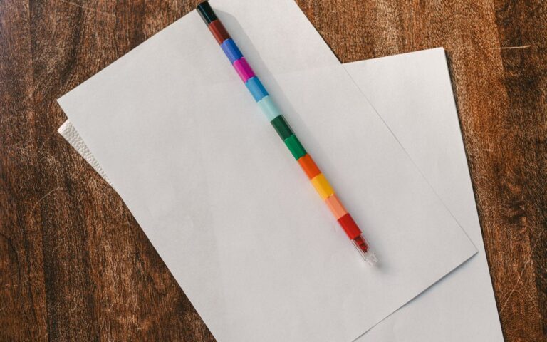 A vibrant multicolored pencil on white blank papers atop a rustic wooden desk.