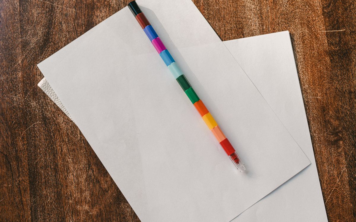 A vibrant multicolored pencil on white blank papers atop a rustic wooden desk.