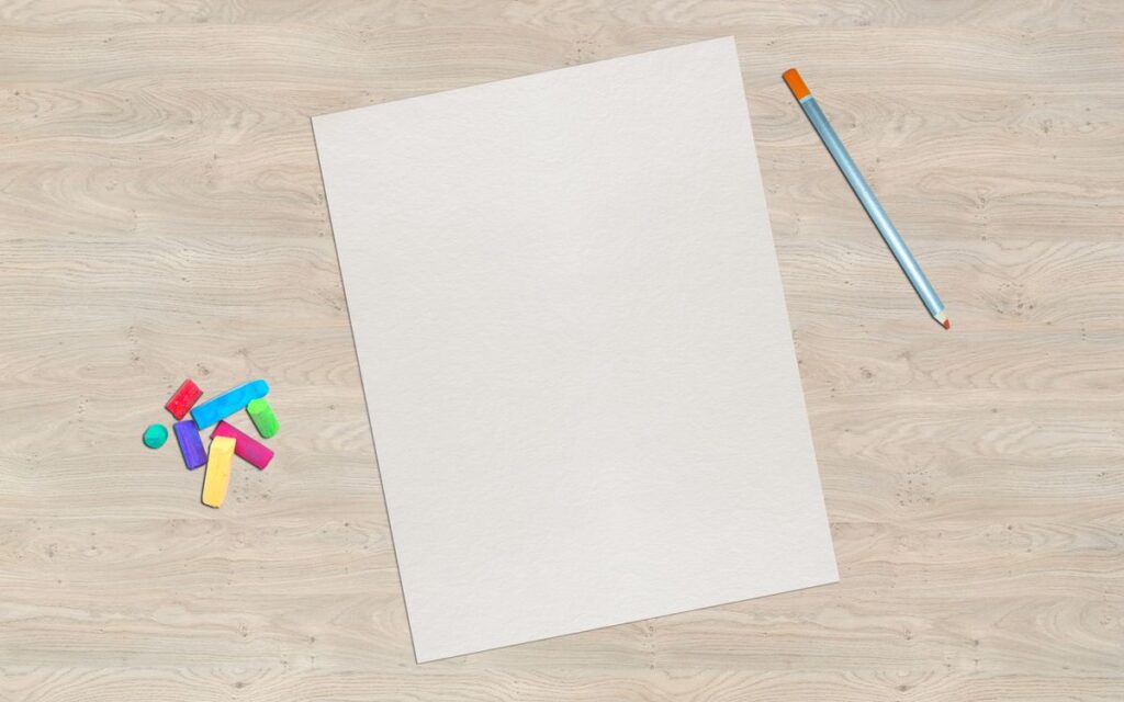 A blank sheet of paper sits on a wooden desk with colorful chalk and a pencil, offering creative possibilities.