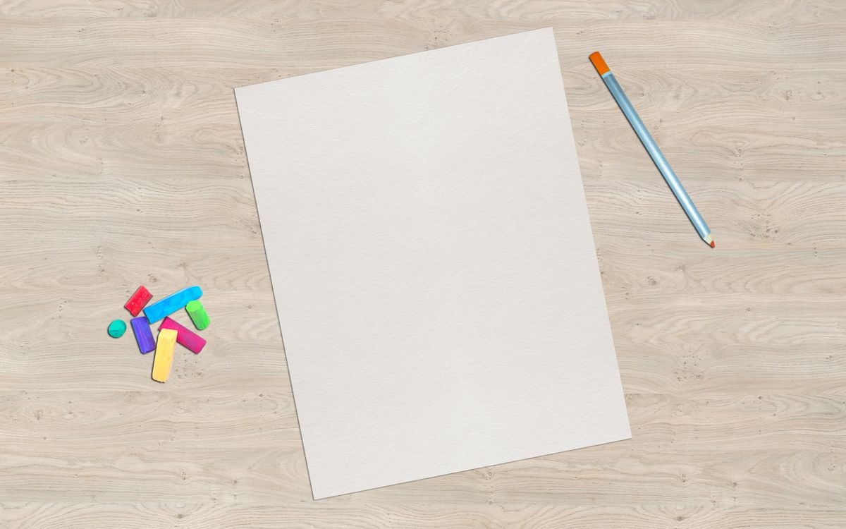A blank sheet of paper sits on a wooden desk with colorful chalk and a pencil, offering creative possibilities.