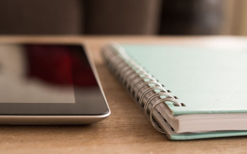 A close-up photo of a tablet alongside a spiral notebook on a wooden desk.