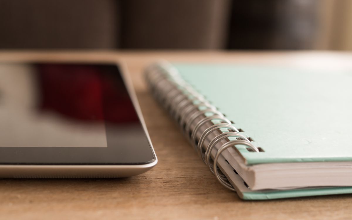 A close-up photo of a tablet alongside a spiral notebook on a wooden desk.