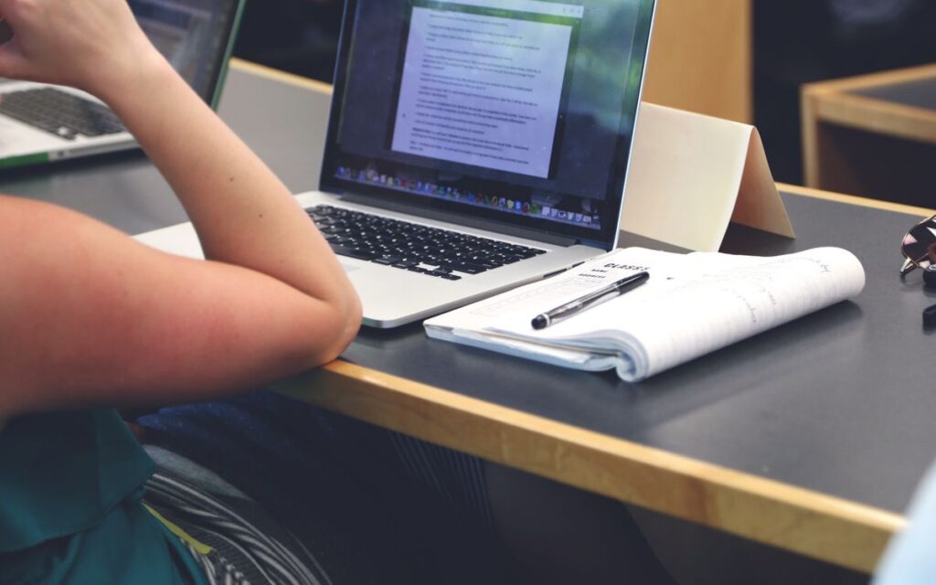 A student in a classroom setting using a laptop and taking notes in a notebook.