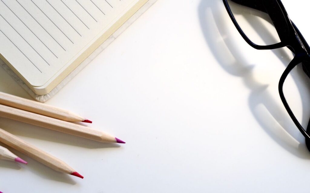 A clean desk setup showing a lined notebook, pencils, and black glasses on a white surface.