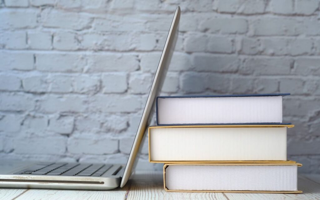 A stack of books next to an open laptop on a wooden desk against a brick wall.