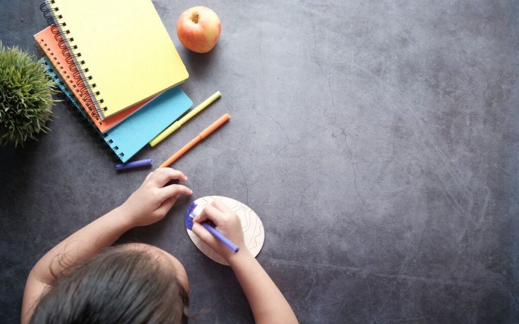 Top view of a child drawing on a textured surface with colorful notebooks and an apple nearby.