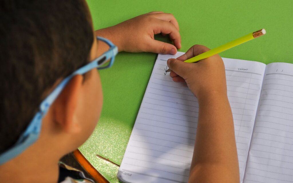 A child wearing glasses writes in a lined notebook with a yellow pencil on a green table.
