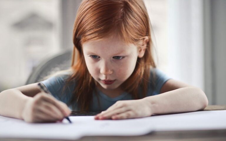 Focused young girl with red hair writing on paper at a desk indoors.