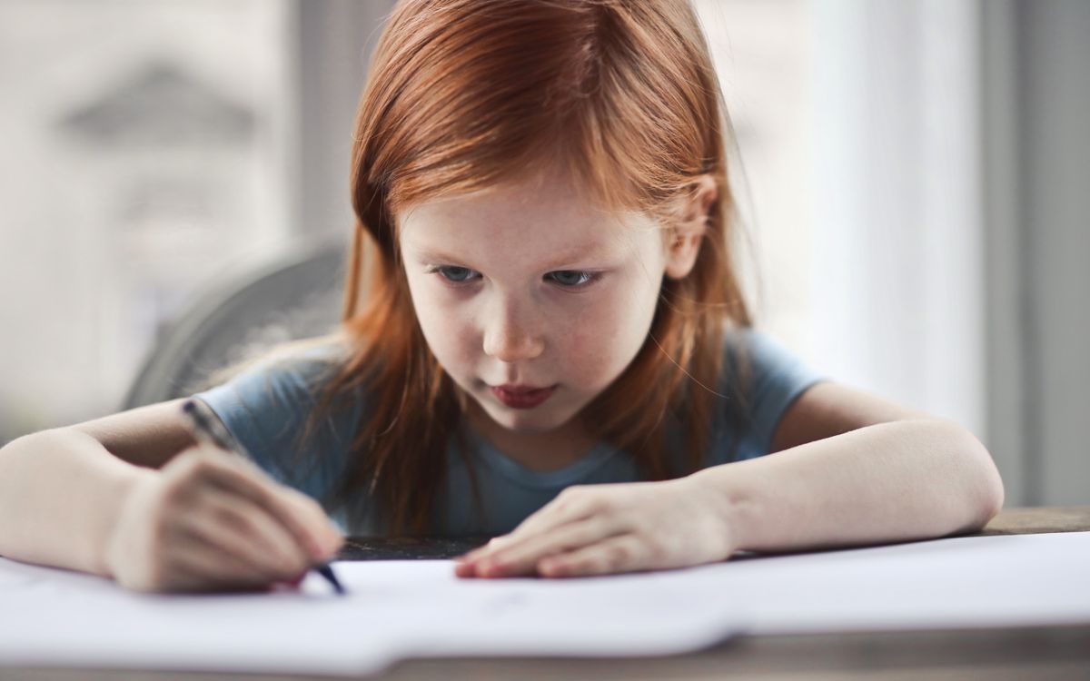 Focused young girl with red hair writing on paper at a desk indoors.