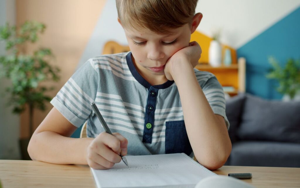 Focused young boy doing homework at a table indoors, deep in thought.