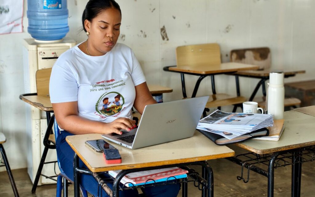 A woman in a classroom typing on a laptop, surrounded by educational materials, conveying a focus on work and study.