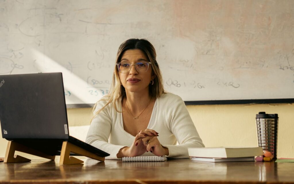 A woman teacher sits at a desk with a laptop and books in a classroom setting.