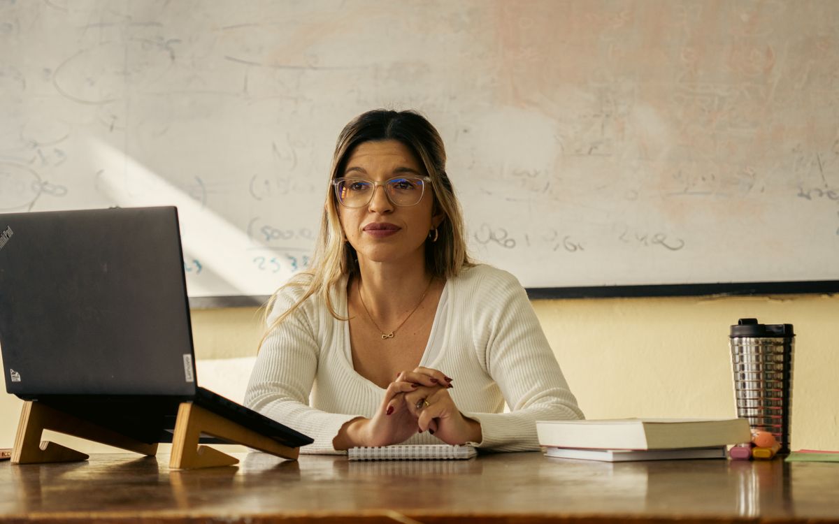 A woman teacher sits at a desk with a laptop and books in a classroom setting.