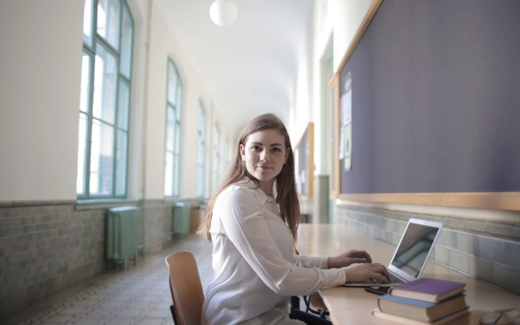 Side view of intelligent young woman in elegant wear sitting at table in university corridor and browsing netbook while