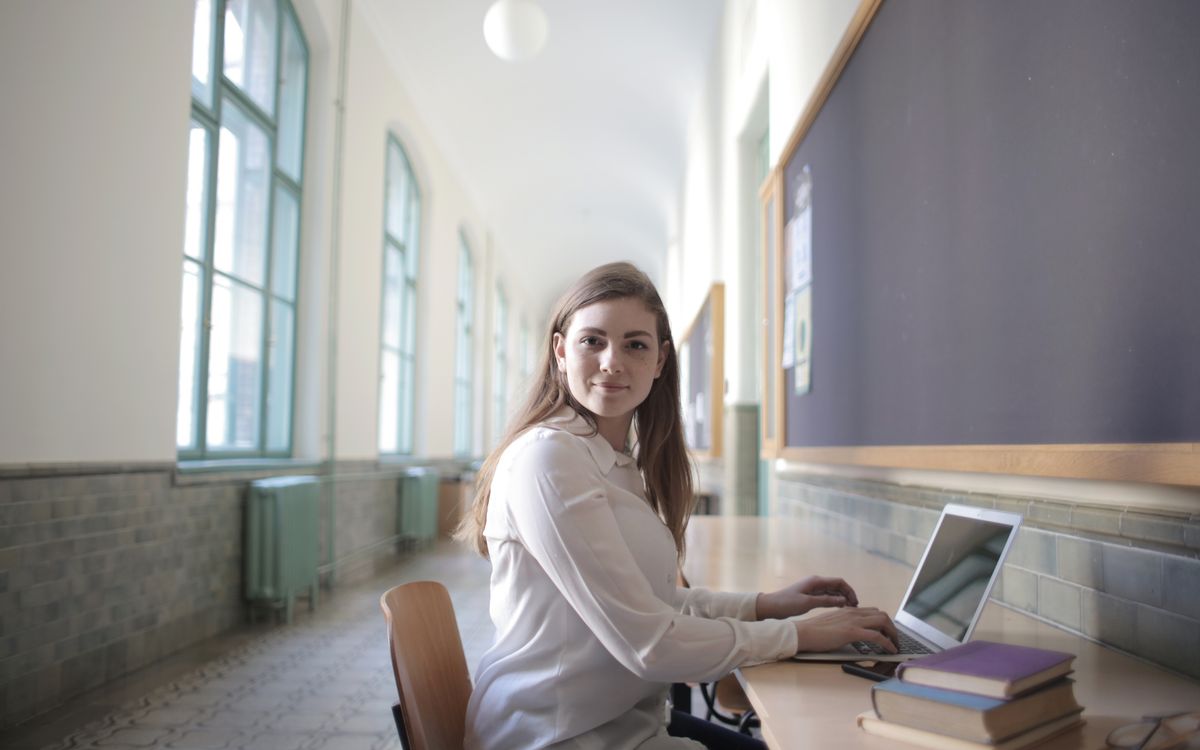 Side view of intelligent young woman in elegant wear sitting at table in university corridor and browsing netbook while