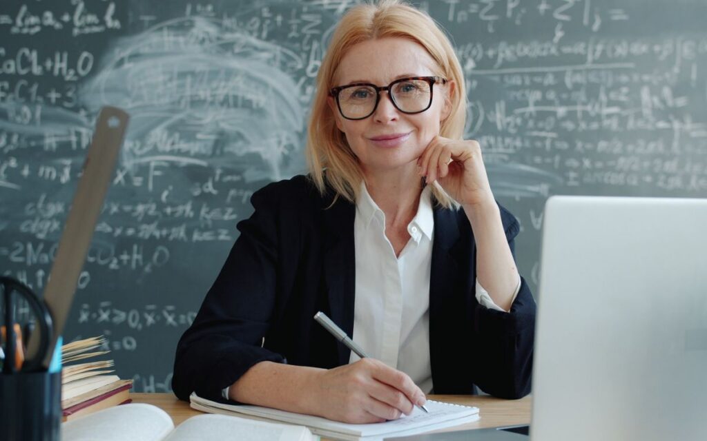 Female teacher smiles at desk with chalkboard in educational setting.