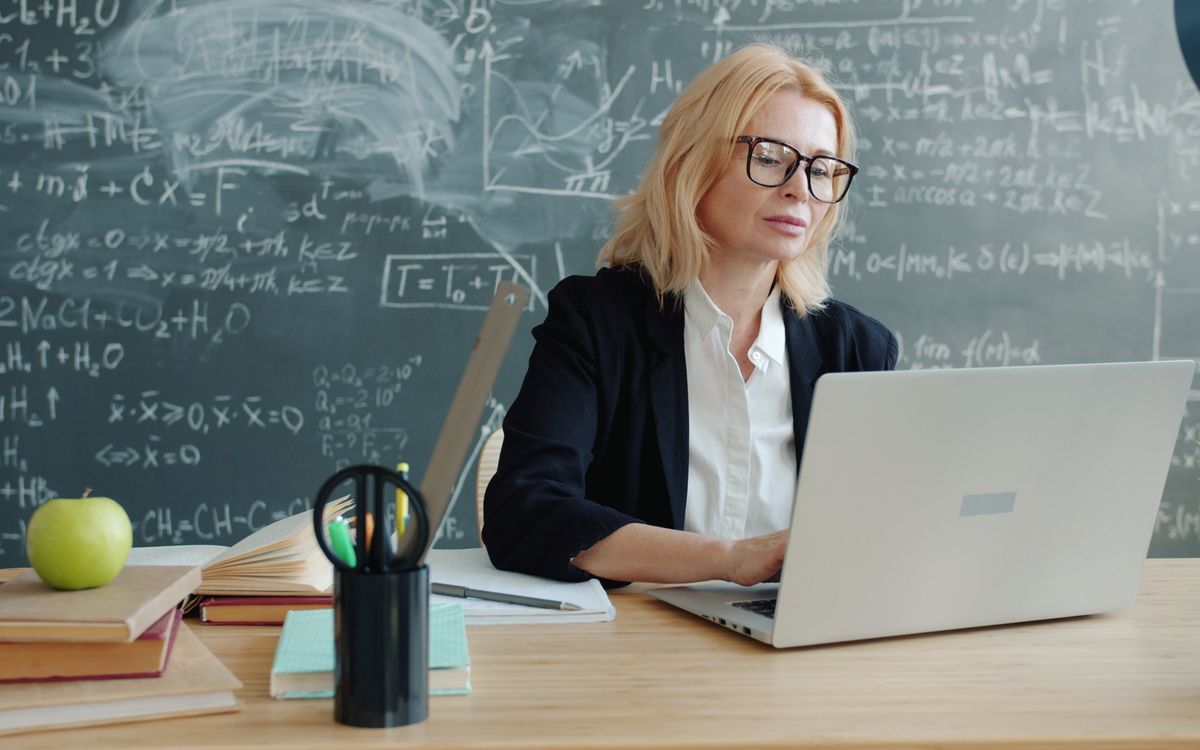 Teacher working on a laptop in a classroom with chalkboard equations, focused and professional.