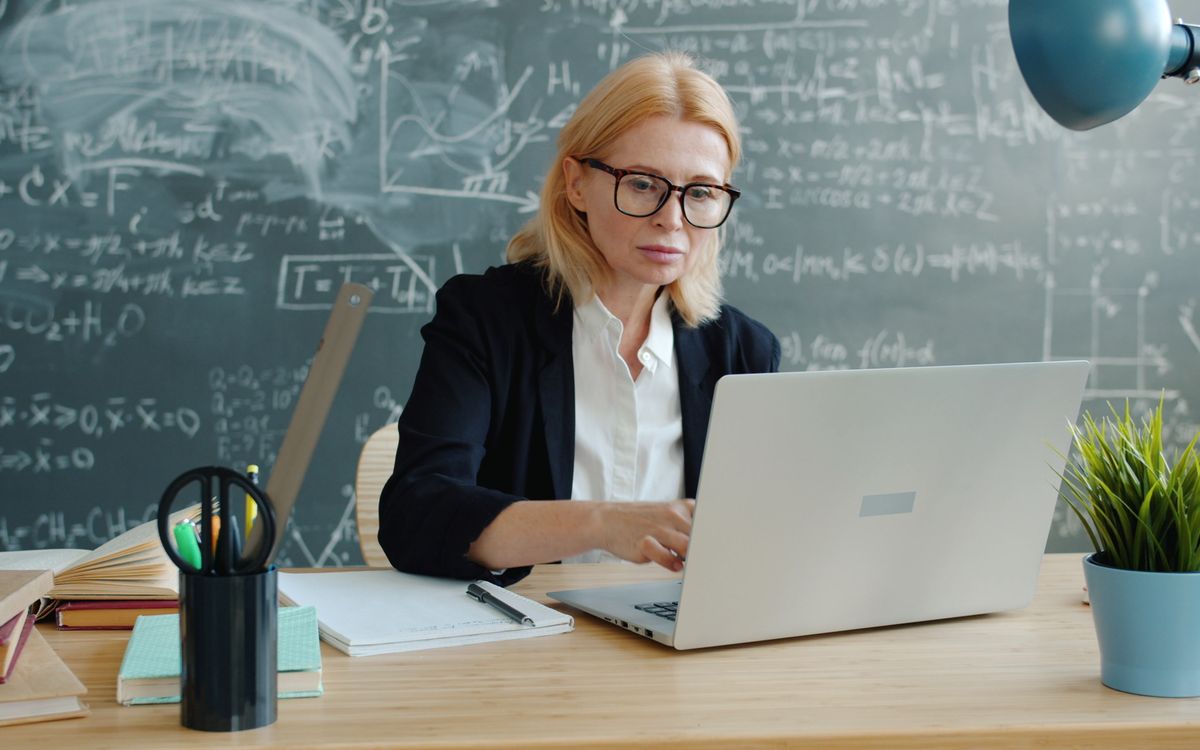 Teacher working on laptop with chalkboard full of equations in background.