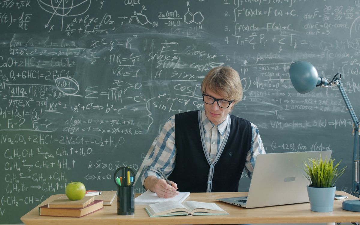 Teacher in classroom writing on notebook, surrounded by books, laptop, and chalkboard.