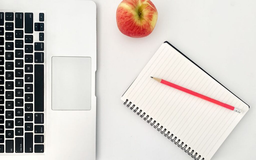 Top view arrangement of modern netbook keyboard placed on white table near opened notebook with blank sheets and red app
