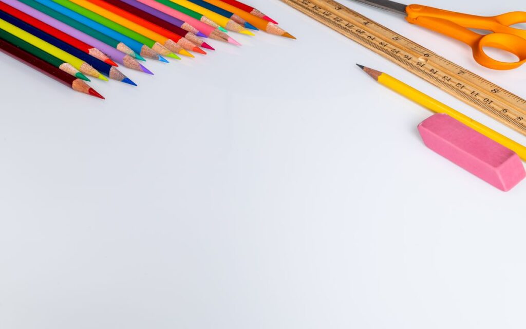 Set of colored pencils for drawing placed on white surface near wooden ruler and scissors near eraser in light studio