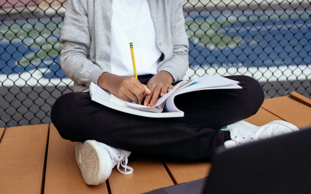 Crop unrecognizable schoolkid sitting with crossed legs and writing notes in copybook while studying with laptop