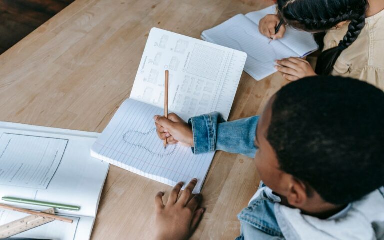 From above of crop unrecognizable students in casual clothes sitting at desk with copybook and doing exercise in classro
