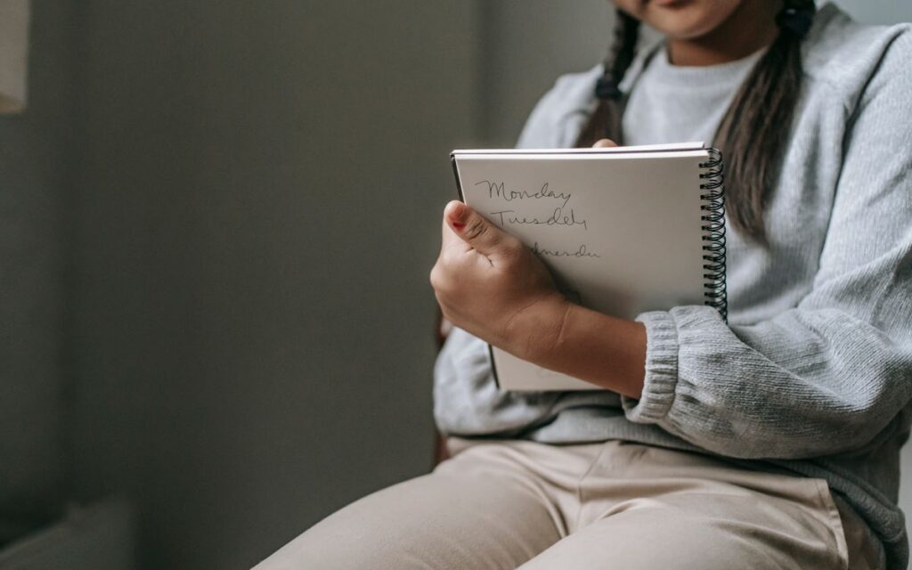 Crop unrecognizable girl in casual outfit sitting on chair and taking notes in copybook