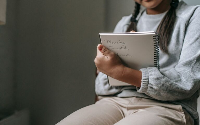 Crop unrecognizable girl in casual outfit sitting on chair and taking notes in copybook