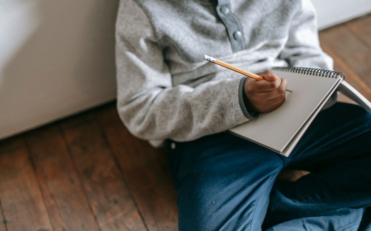 From above faceless child with pencil and notebook taking notes while sitting on floor