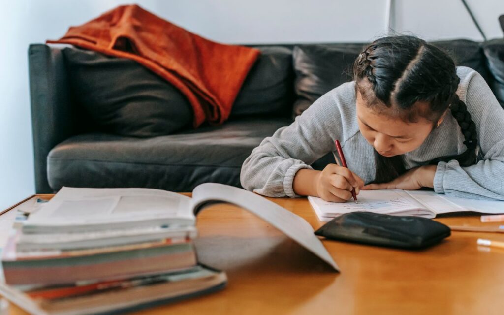 Attentive Asian schoolchild writing in workbook with pen while doing homework at table at home