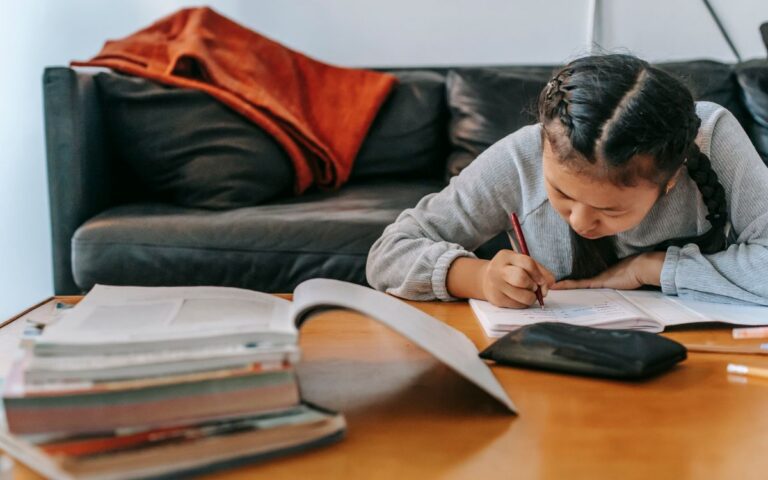 Attentive Asian schoolchild writing in workbook with pen while doing homework at table at home