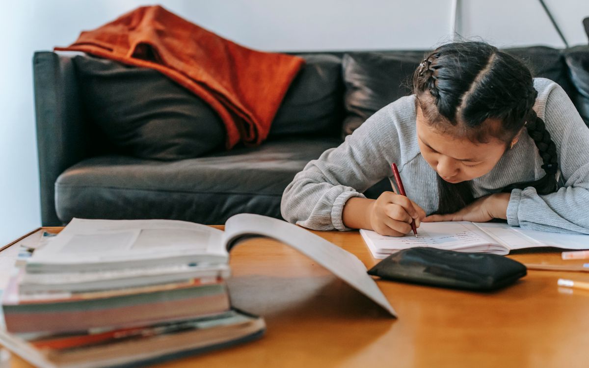 Attentive Asian schoolchild writing in workbook with pen while doing homework at table at home