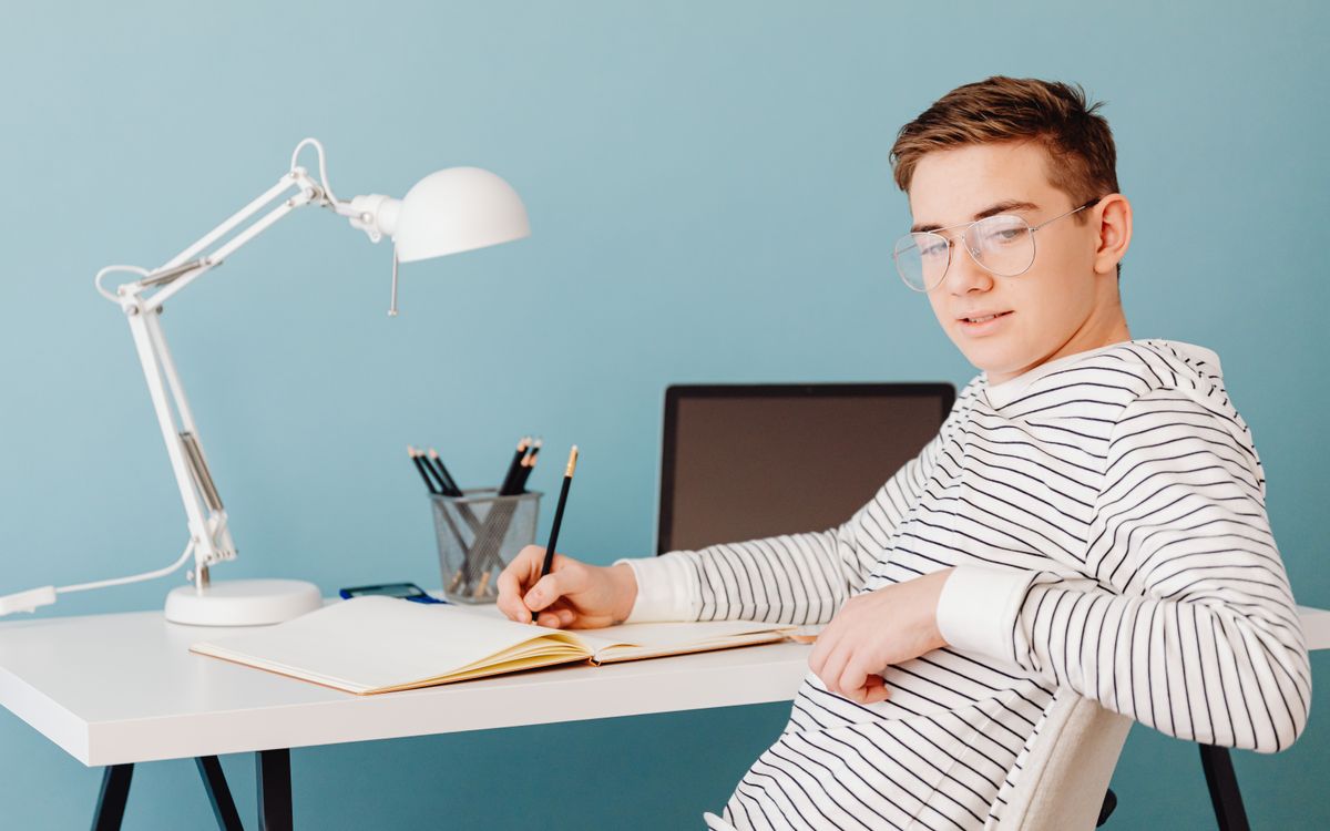 Young male student wearing glasses studying at a desk with a laptop and lamp.