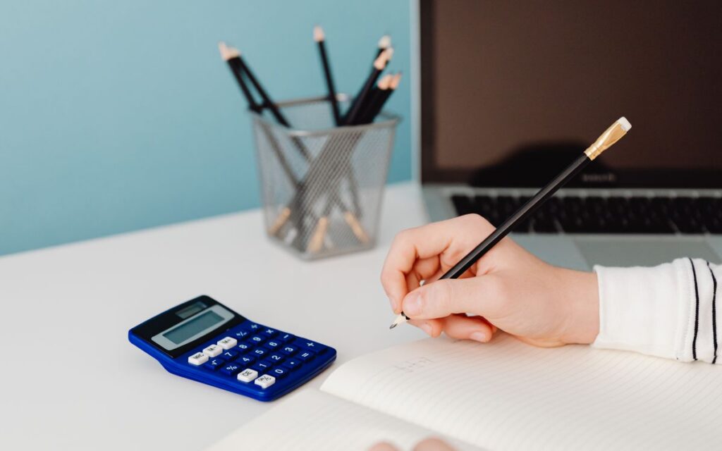 Close-up of a hand writing in a notebook beside a calculator and laptop on a desk.