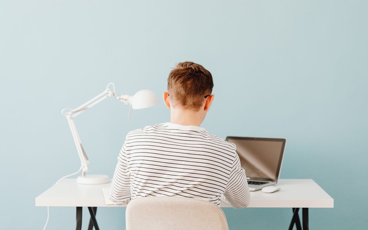 Teenager using laptop at desk under lamp in a minimalist setting with blue background.