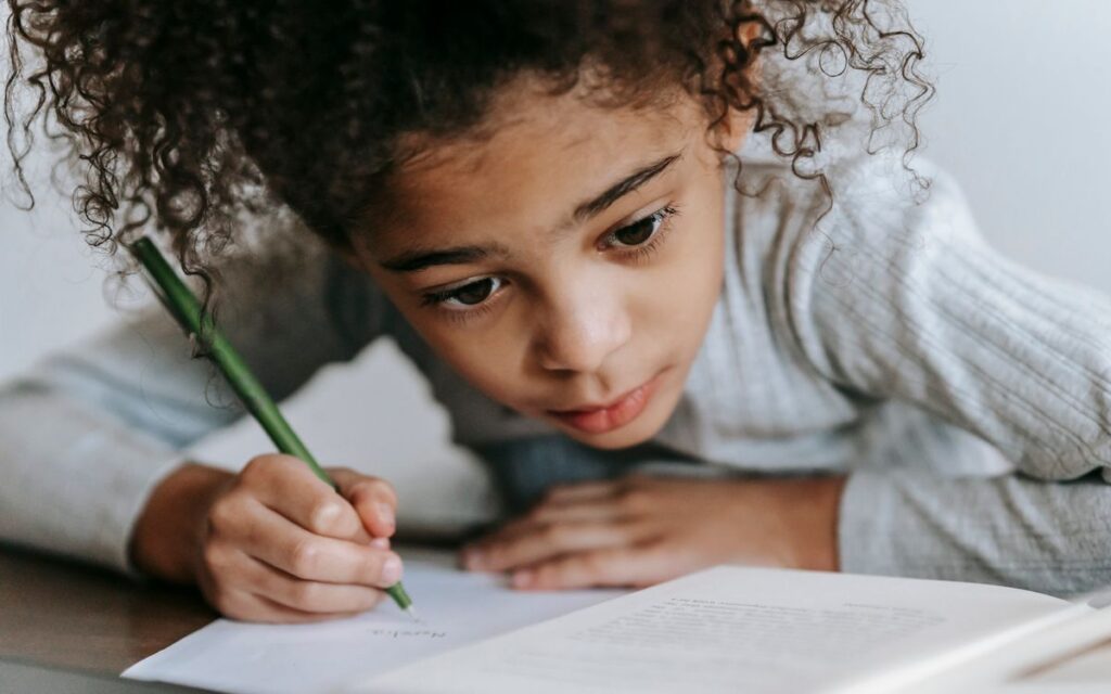 Crop concentrated African American girl with curly hair sitting at table with textbook while taking notes on sheet of pa