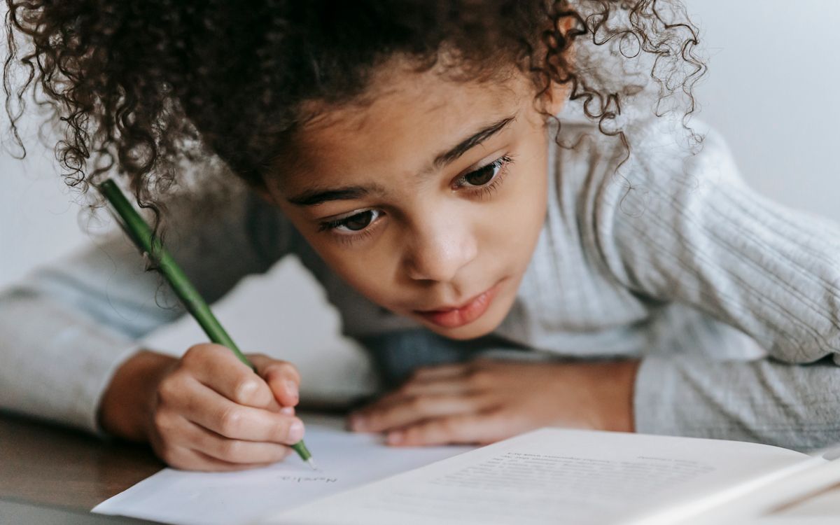 Crop concentrated African American girl with curly hair sitting at table with textbook while taking notes on sheet of pa