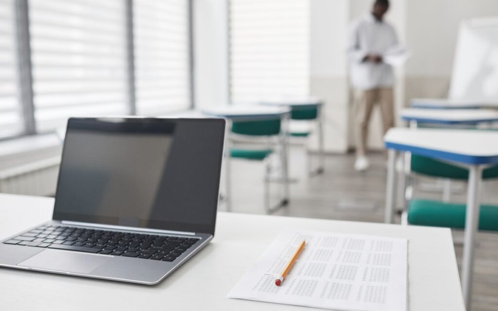 Laptop and exam sheet on a desk in a modern classroom setting.