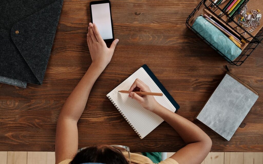 Overhead view of a child writing in a notebook while using a smartphone at a wooden desk.