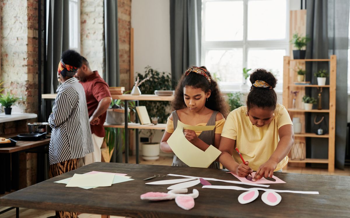 Two children crafting with paper indoors while adults cook in the background.