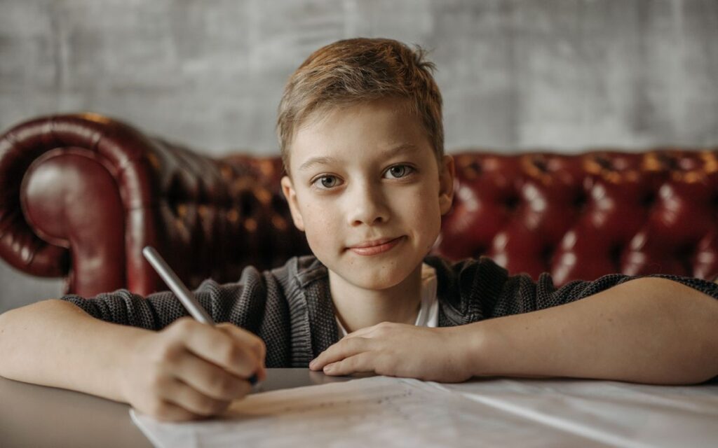 Smiling young boy writing on paper seated comfortably on a leather sofa in a cozy indoor setting.