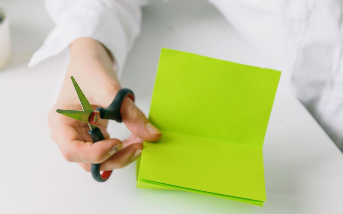 Close-up of a hand holding scissors while cutting bright green paper indoors.