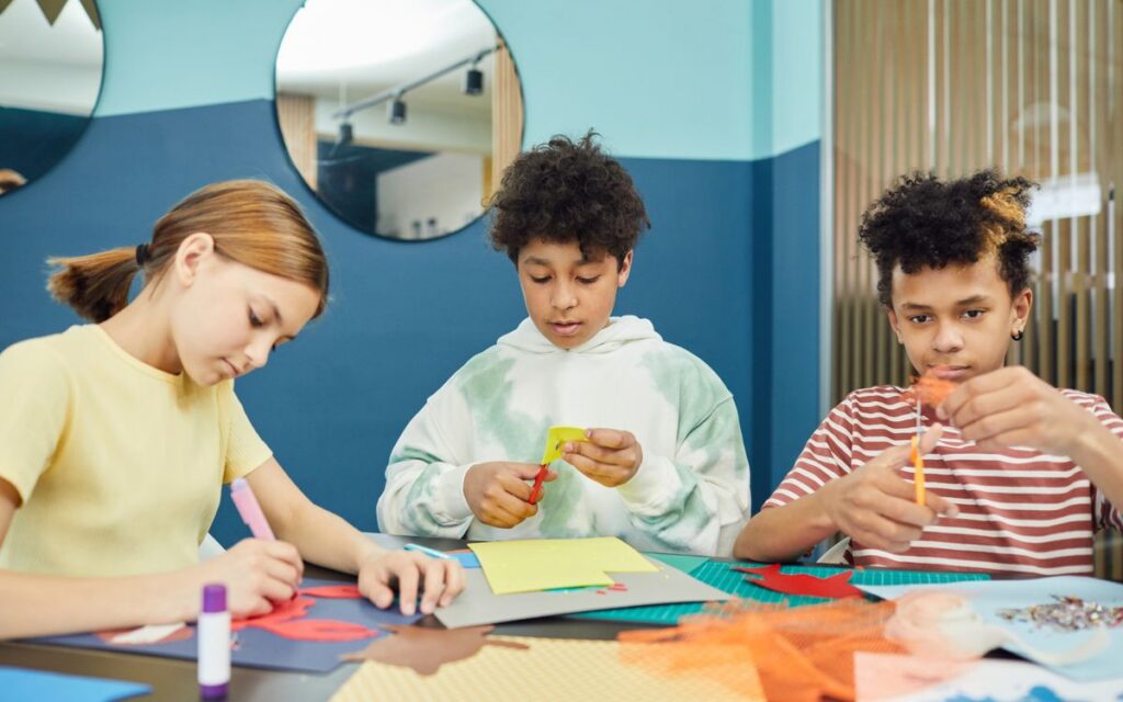 Group of concentrated multiracial children making artworks with colorful paper and scissors while sitting at table in li