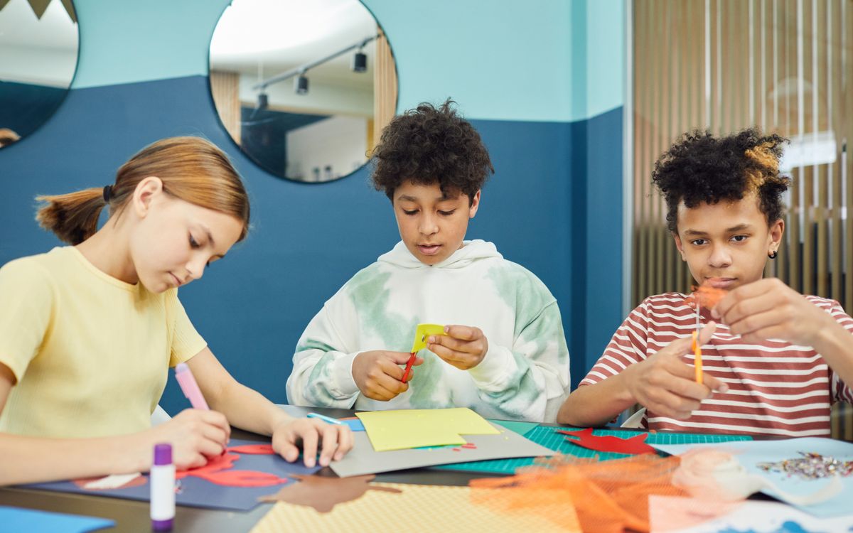 Group of concentrated multiracial children making artworks with colorful paper and scissors while sitting at table in li